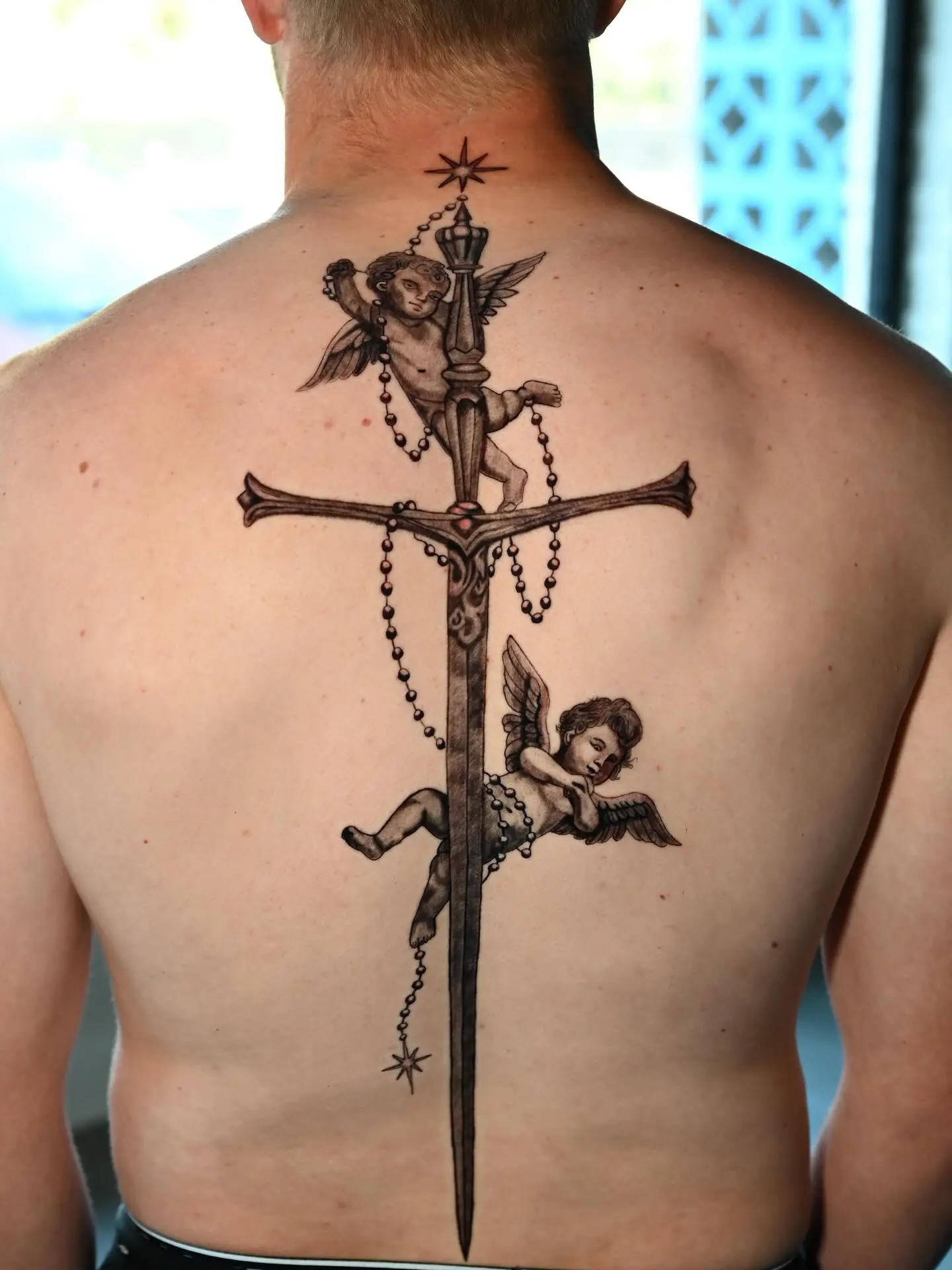 Black and grey back tattoo of two cherubs holding a sword and rosary by Gynmo at Shadow Studio Charlotte