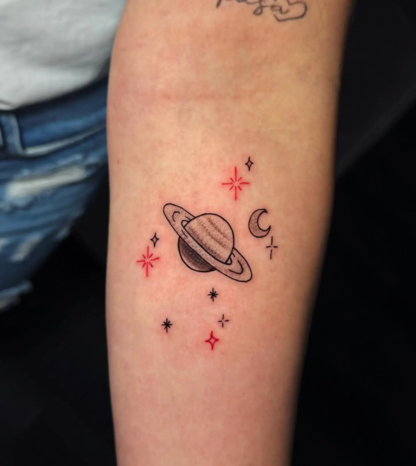 Fine line forearm tattoo of Saturn, moon, and stars with red sparkle accents by Venus at Shadow Studio Charlotte